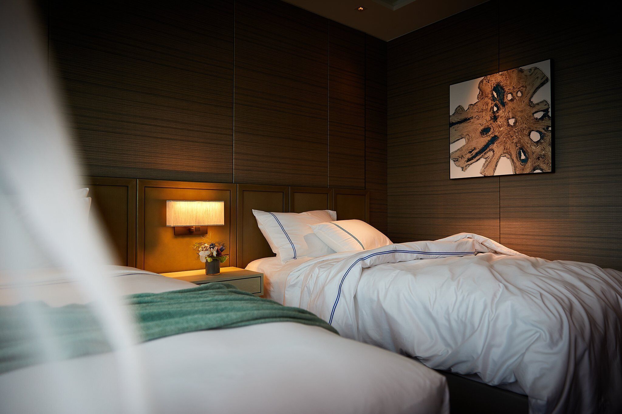 Quarto de hotel com duas camas, uma mesa de cabeceira com abajur e uma obra de arte na parede.