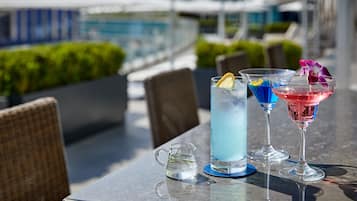 Un tavolo con tre bevande e una caraffa d'acqua, all'aperto con vista su una piscina e vegetazione.