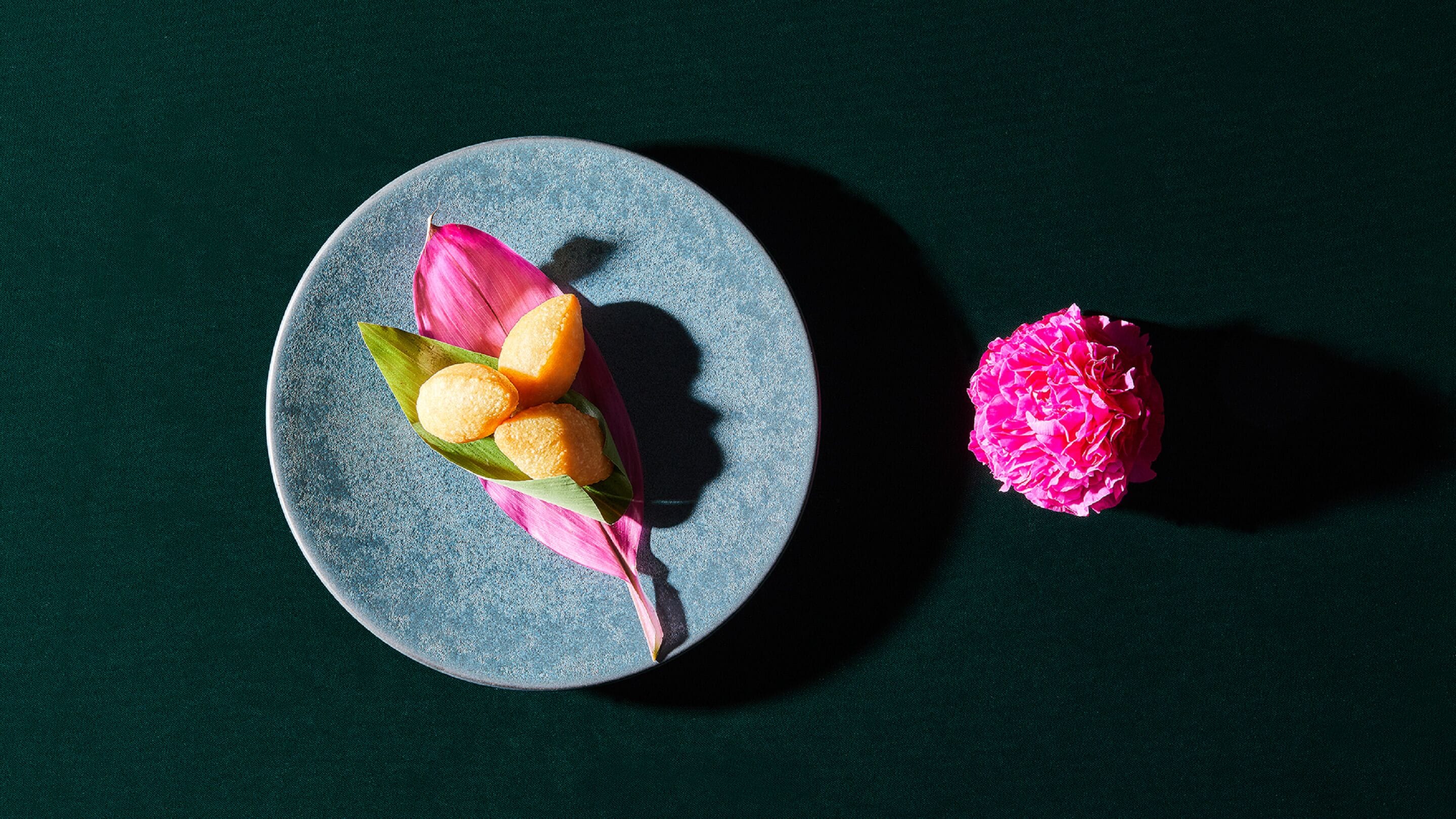 Un plato con tres piezas redondas y de color marrón dorado sobre una hoja verde, y una flor rosa sobre una superficie oscura.