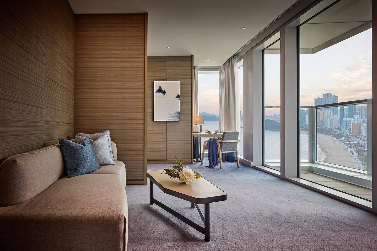 A modern hotel room with a large window offering a city view, a sofa with cushions, a small table with a flower arrangement, and a desk with a lamp.