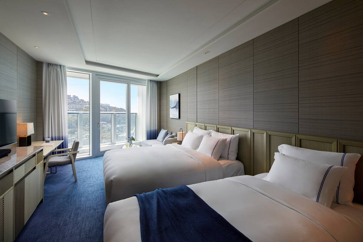 A hotel room with two beds, a desk, a chair, and a large window offering a city view.