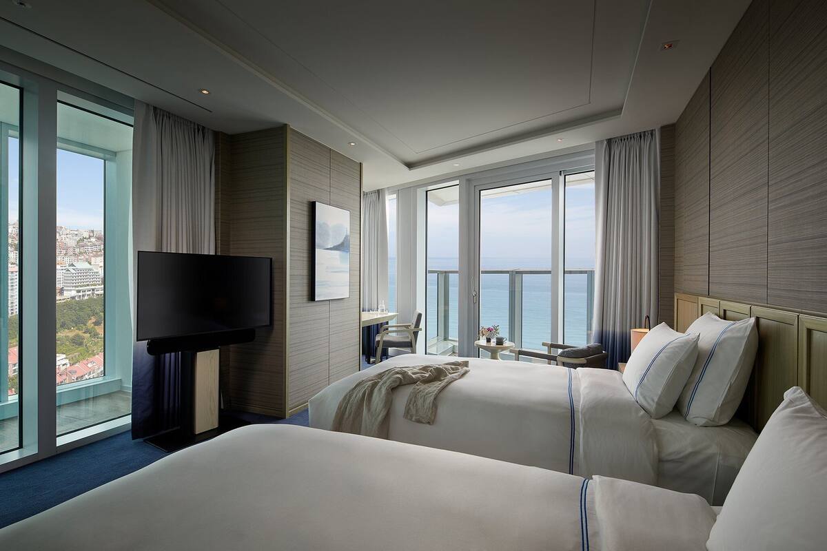 A hotel room with two beds, a TV, a desk, and a view of the city and ocean.