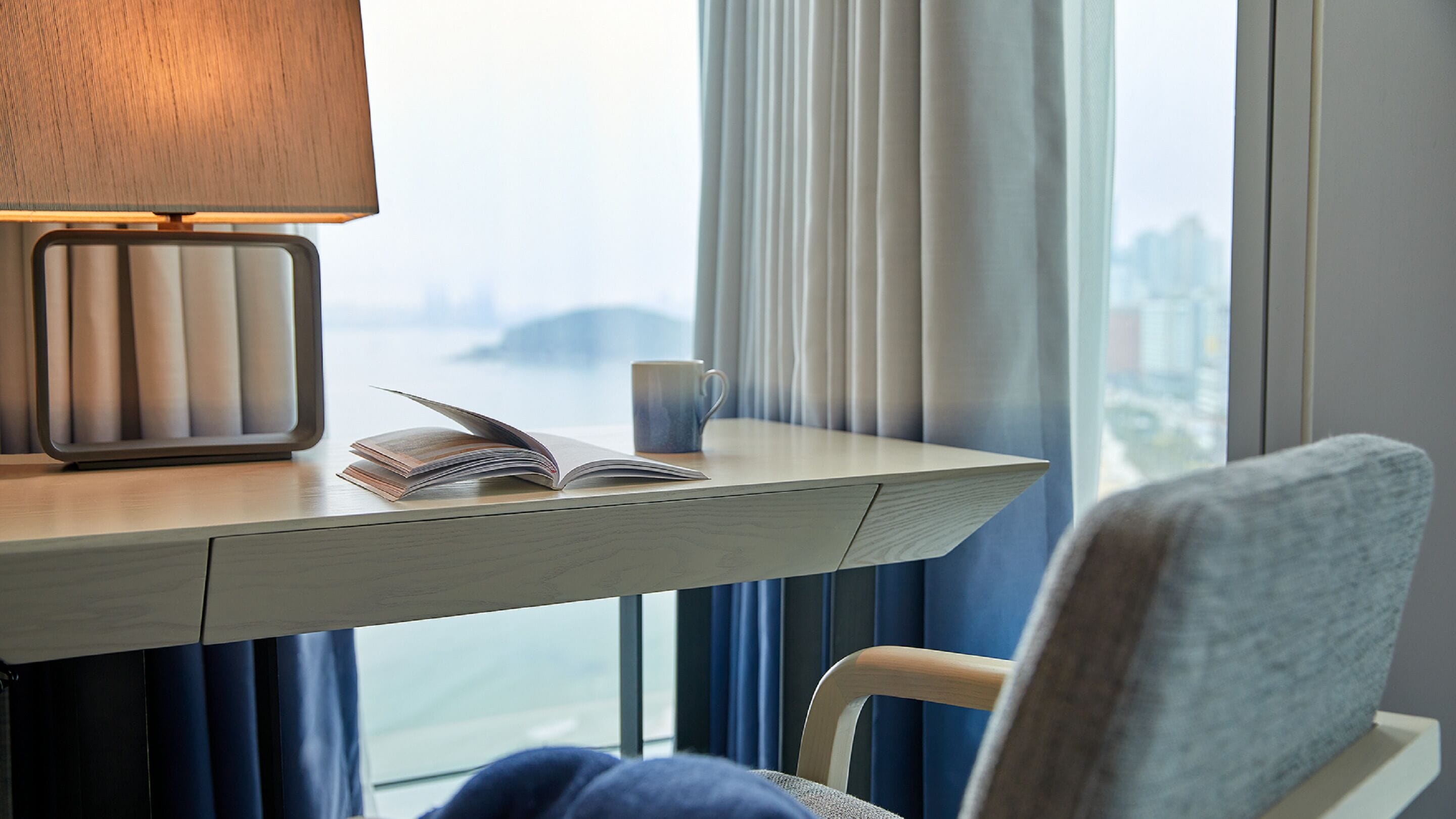 A hotel room with a bedside table, a lamp, an open book, a mug, and a view of a cityscape through the window.