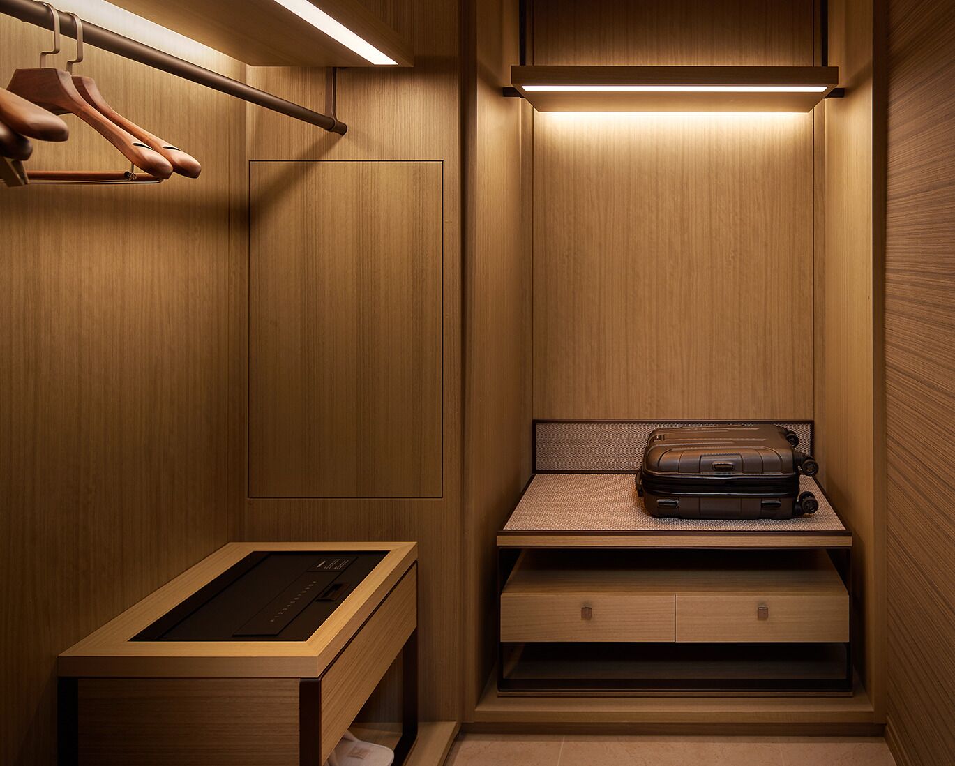 Ein kleiner, gut beleuchteter begehbarer Kleiderschrank mit Holzwänden, einer Kleiderstange, einer Sitzbank mit Schubladen und einem Koffer auf der Bank.