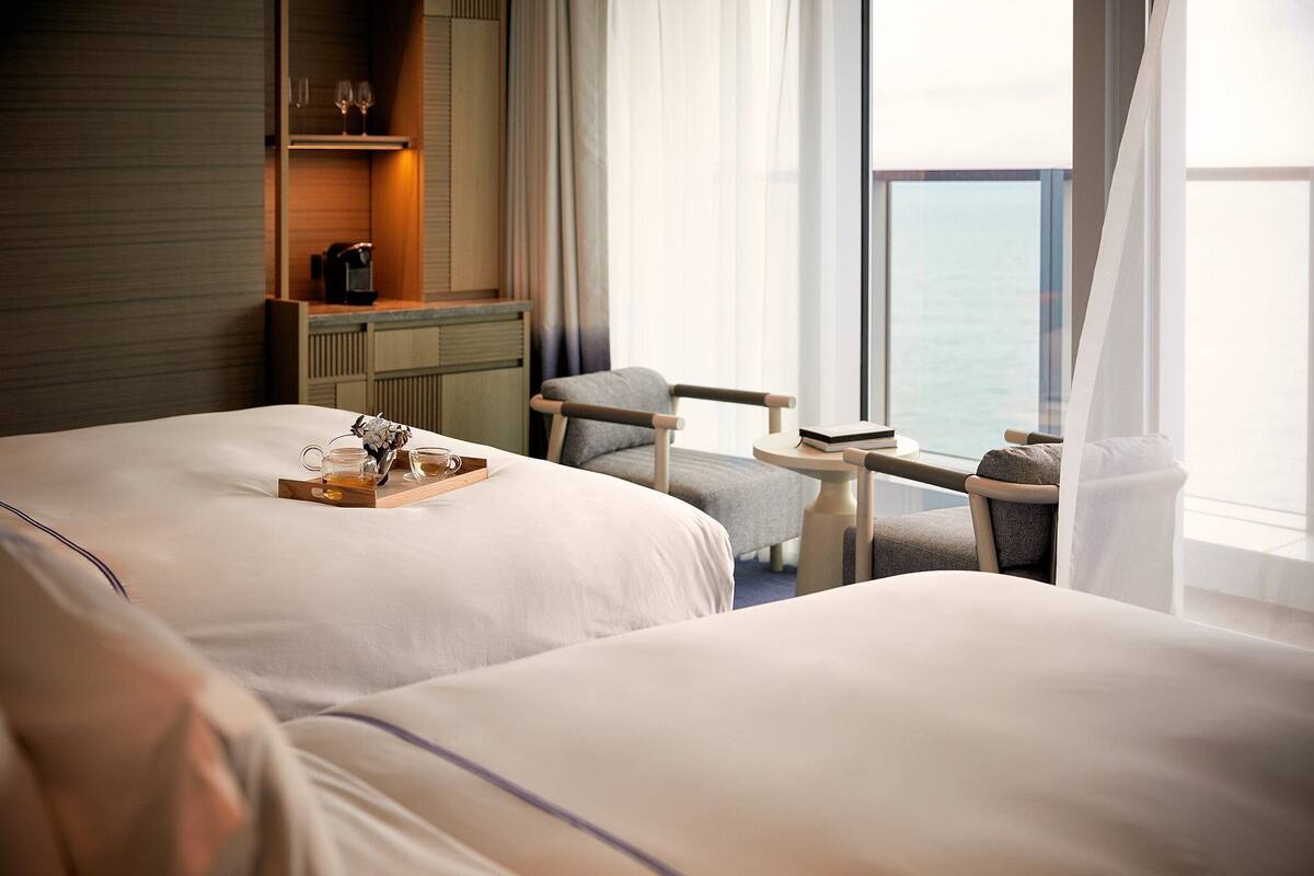 A hotel room with two beds, a small table with a tray of tea, a chair, and a view of the sea through large windows.