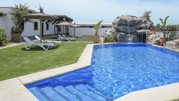 Ein Schwimmbecken mit Felsen, weiße Liegestühle und ein kleines Haus mit einem Strohdach.
