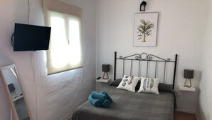 Ein Schlafzimmer mit Bett, Wandfernseher, Fenster, Nachttischlampe und einer gerahmten Pflanze.