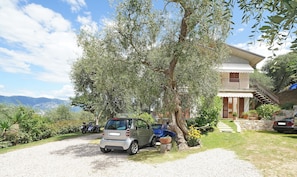 A house with a gravel driveway, parked cars, and a large tree in front. - Apartment "Anna Torri" with Beautiful Garden and Lake Views (Torri del Benaco)