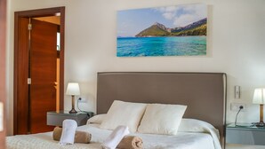 Habitación de hotel con una cama, dos mesitas de noche, un cuadro de un paisaje costero y una puerta.