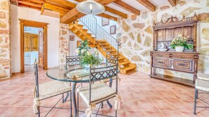 Amplia habitación con pared de piedra, vigas de madera en el techo, mesa de vidrio con sillas, una escalera y un mueble de madera con objetos decorativos.