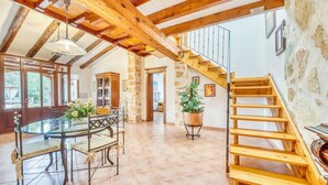 Una habitación amplia con una mesa de comedor de cristal, vigas de madera, una escalera y vistas al exterior.