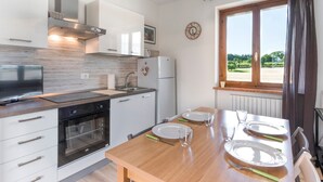 A modern kitchen with white cabinets, a built-in oven, a dining table set for four, and a view of a green landscape through a window. - Holiday Apartment "Bella Cora" With Garden and Private Rooftop Terrace (LAZISE)