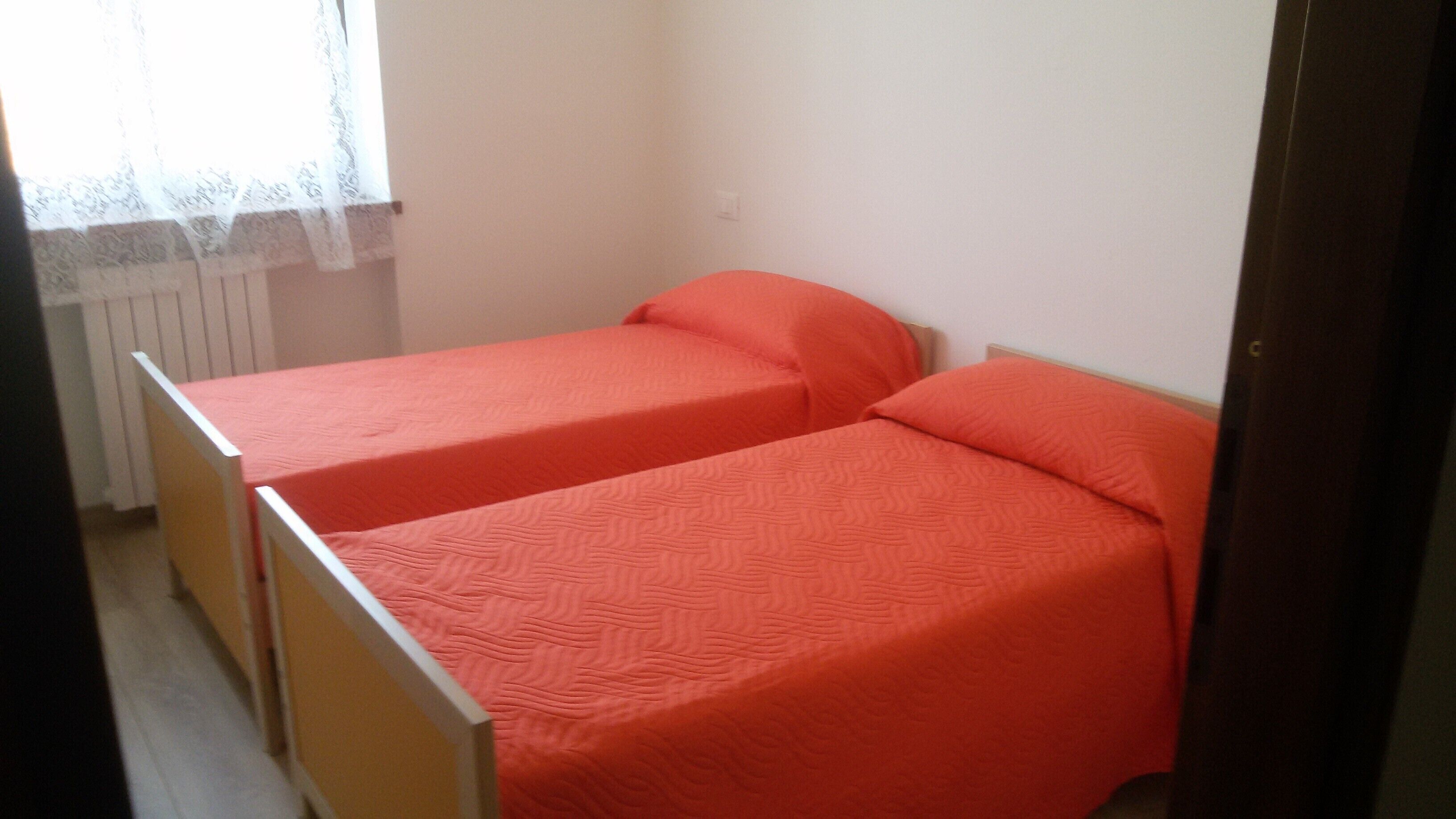 Una habitación pequeña con dos camas individuales, un radiador y una ventana con cortina translúcida.