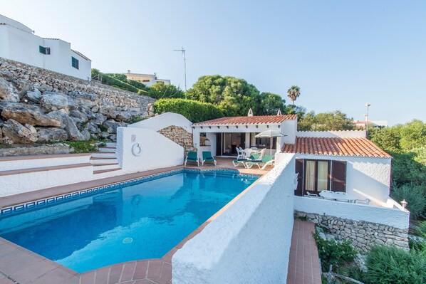 A swimming pool with a stone border, a white building with a tiled roof, and a stone wall.