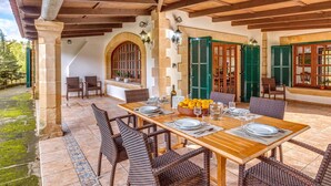 Un espacio comedor al aire libre con una mesa de madera dispuesta para cuatro personas, rodeada de sillas de mimbre, bajo un techo de madera con vigas a la vista.