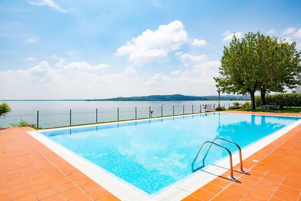 A rectangular swimming pool with clear blue water, surrounded by a tiled deck, a metal fence, and a tree.