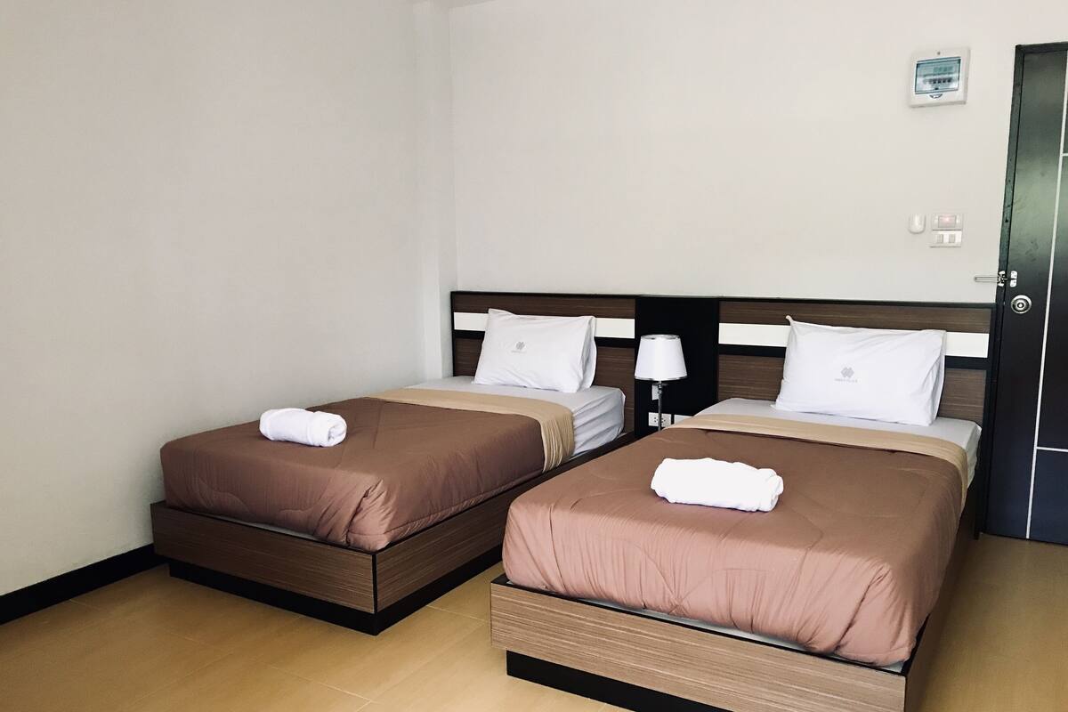 A hotel room with two beds, each with a brown bedspread and white pillows, a bedside table with a lamp, and a wall-mounted thermostat.