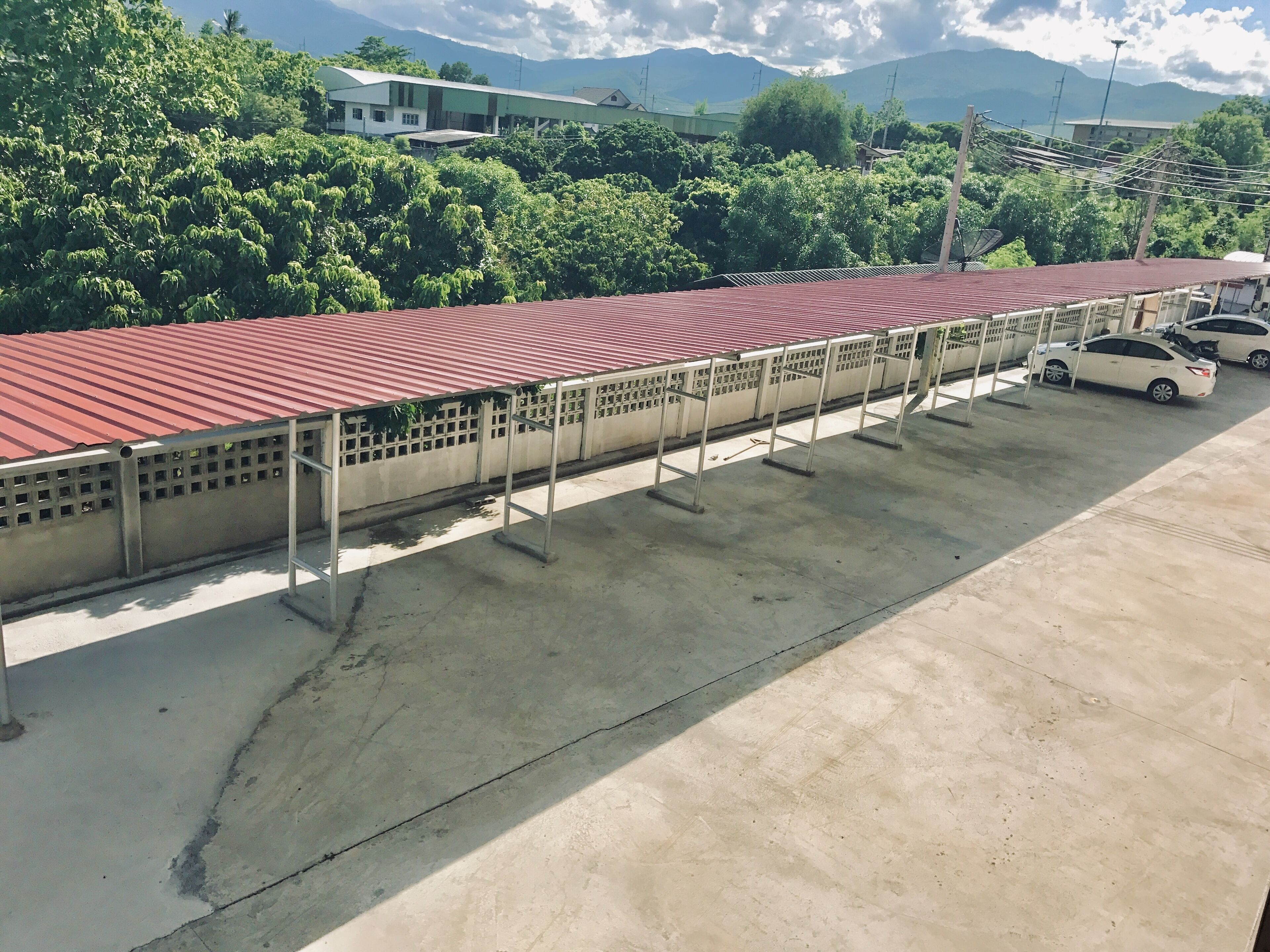 Un estacionamiento con techo rojo y suelo de concreto, rodeado de vegetación y edificios.