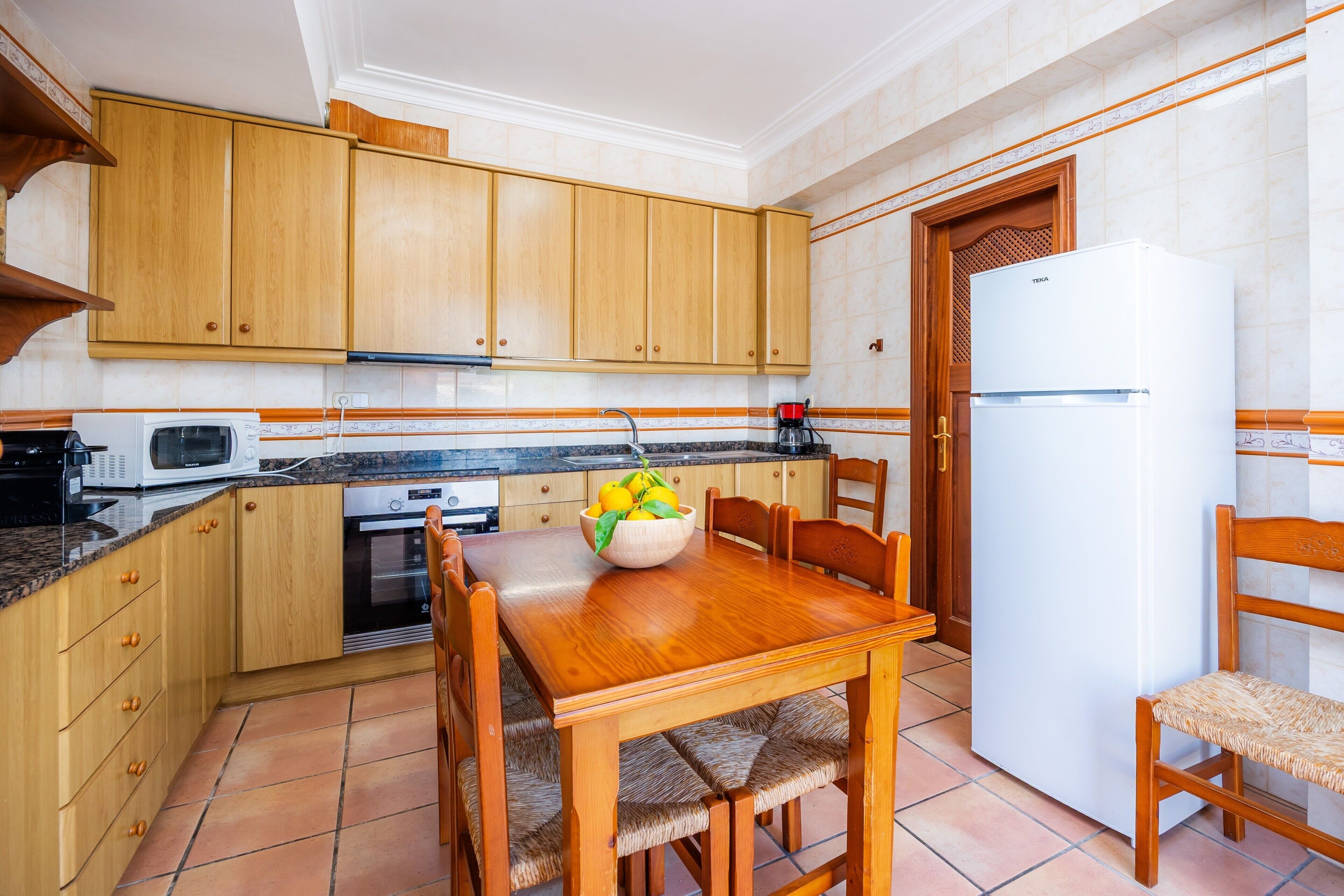 Una cocina con armarios de madera, una mesa de comedor, sillas, un refrigerador y un microondas.