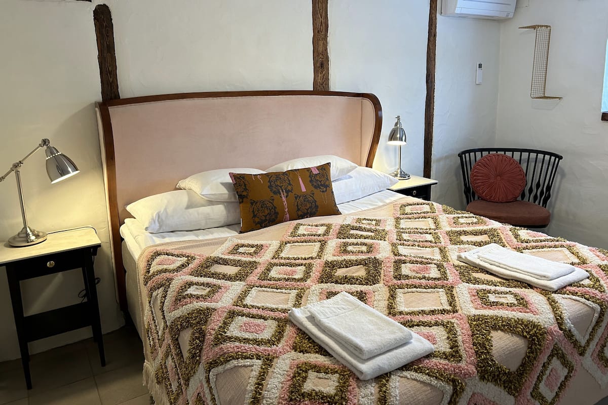Uma cama bem arrumada com um cobertor estampado, uma luminária de cabeceira, uma cadeira e um aparelho de ar condicionado na parede.