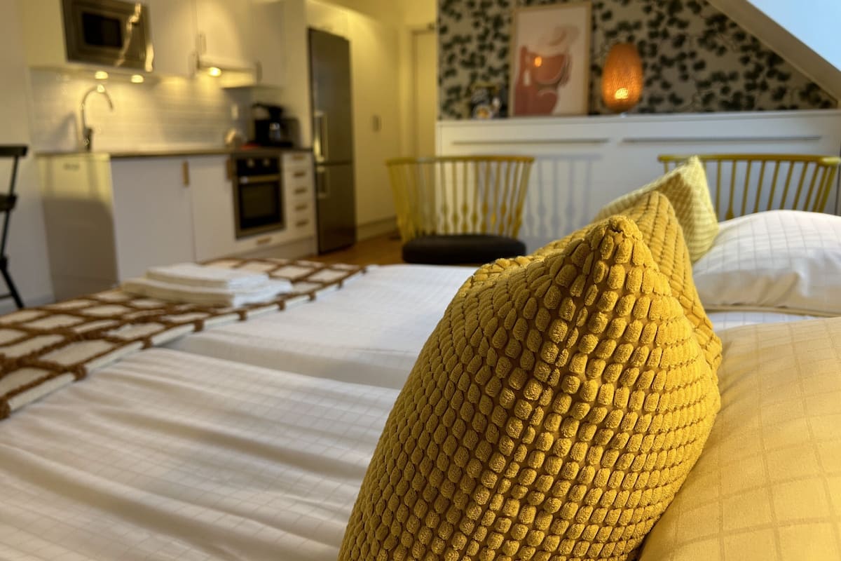 Quarto com cama, almofadas e vista para uma cozinha e varanda.