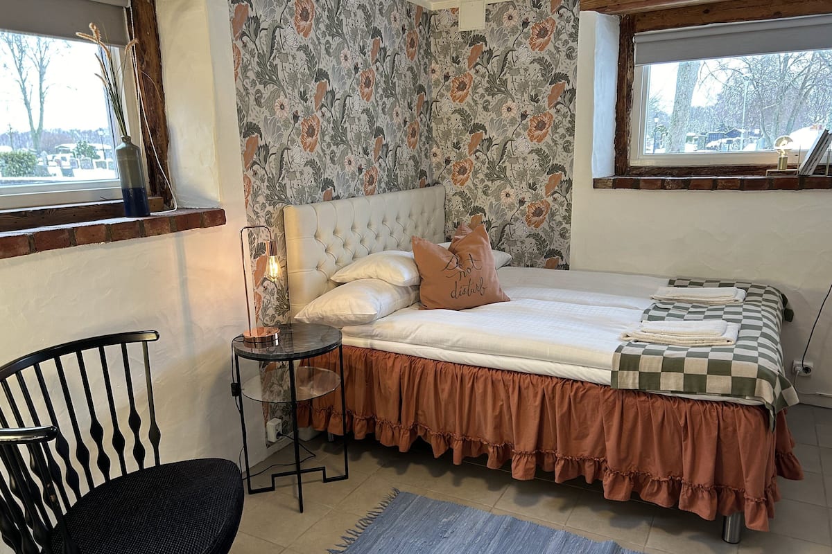 Quarto com cama, cadeira, mesinha e janela.