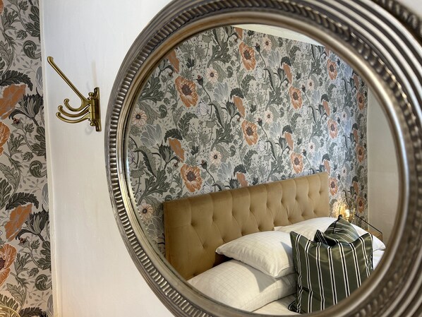 A bedroom with a tufted headboard, floral wallpaper, a brass wall sconce, and a striped pillow on a nightstand. - Halmstad Gårdshotell (Halmstad)