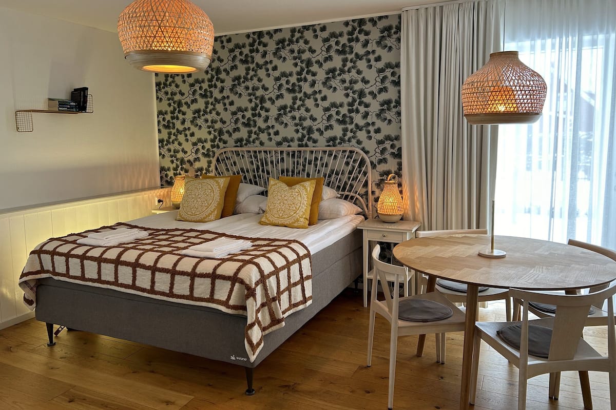 Quarto com cama, luminárias de cabeceira, uma mesa redonda com cadeiras e uma janela com cortinas.