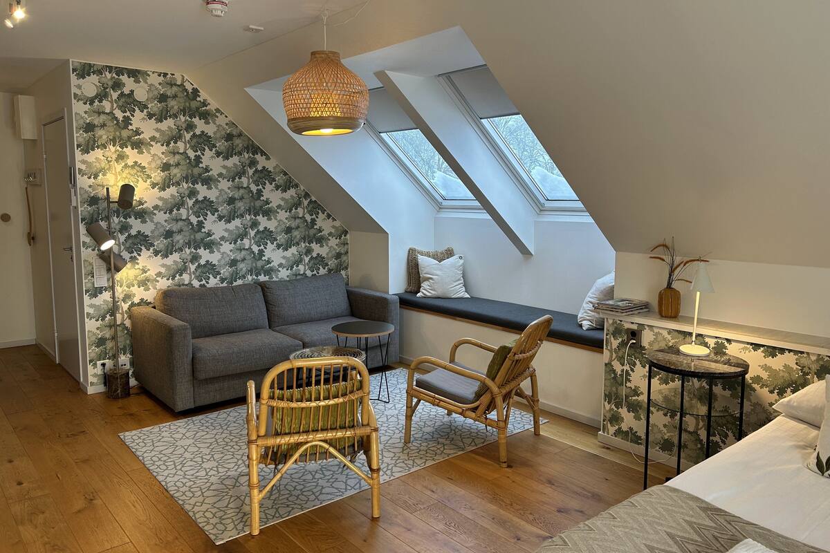 Um quarto de loft aconchegante com teto inclinado, um sofá cinza, duas cadeiras de vime, uma mesa redonda pequena e um assento de janela com vista para o céu.