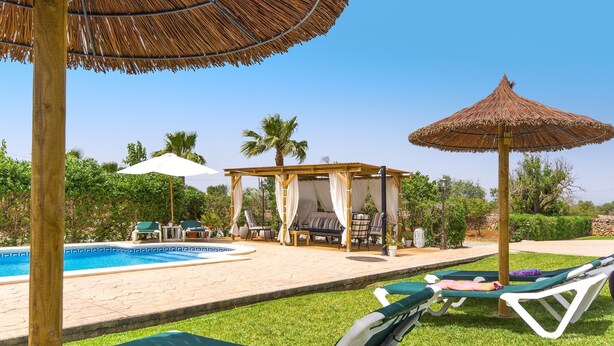 Un espace piscine avec des chaises longues, un coin salon abrité et un parasol en chaume.
