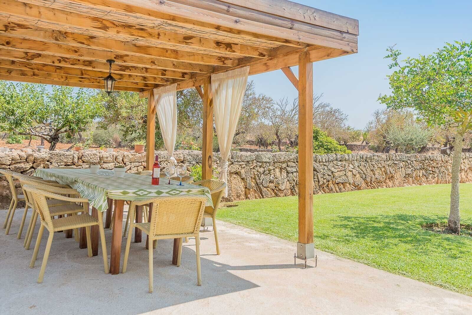 En udendørs patio med et bord og stole, en pergola over hovedet og en stenvæg i baggrunden.