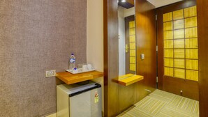 A hotel room with a wooden door, a small refrigerator, a wooden shelf with a bottle and glasses, and a patterned floor. - The Ashoka Hotel (Indore)