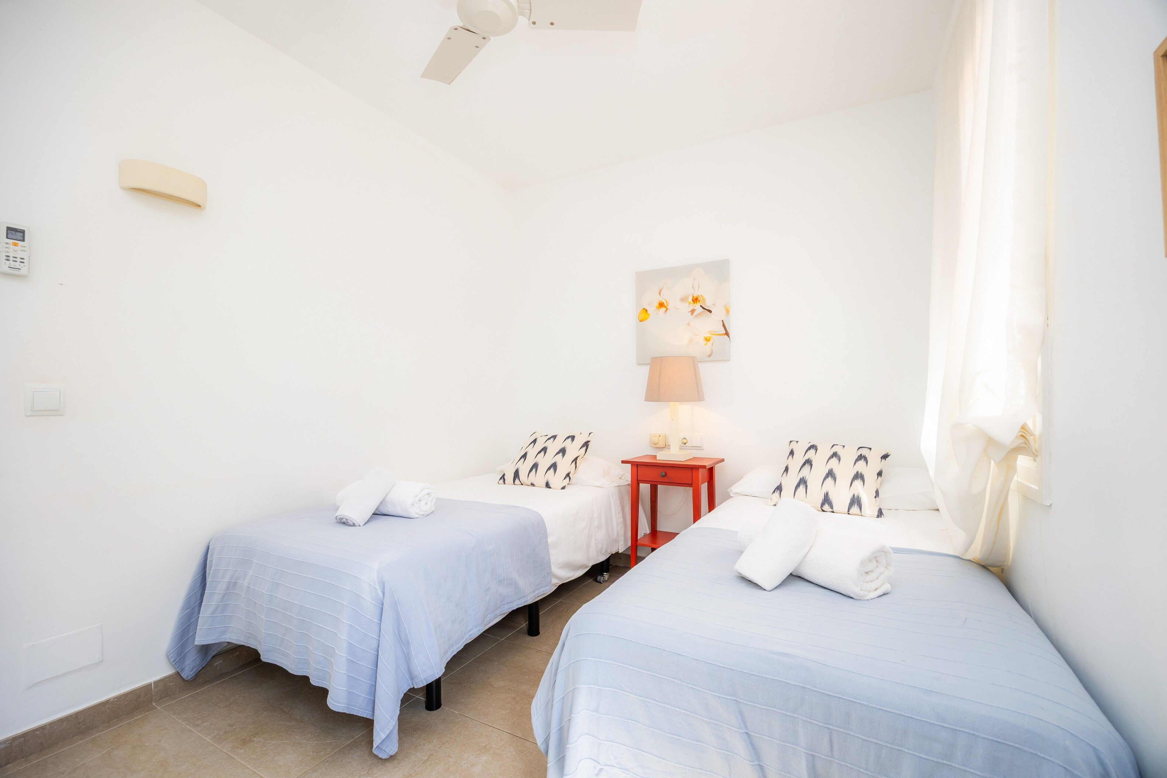 Une chambre d’hôtel avec deux lits, une table de chevet, un ventilateur de plafond, un téléphone fixé au mur et un tableau.