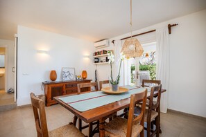 A dining area with a wooden table, chairs, a hanging light fixture, and a view of greenery outside. - Beautiful "Can Botana 7" with Wi-Fi, Pool, Air Conditioning, Balcony and Garden (Cala Sant Vicenç)