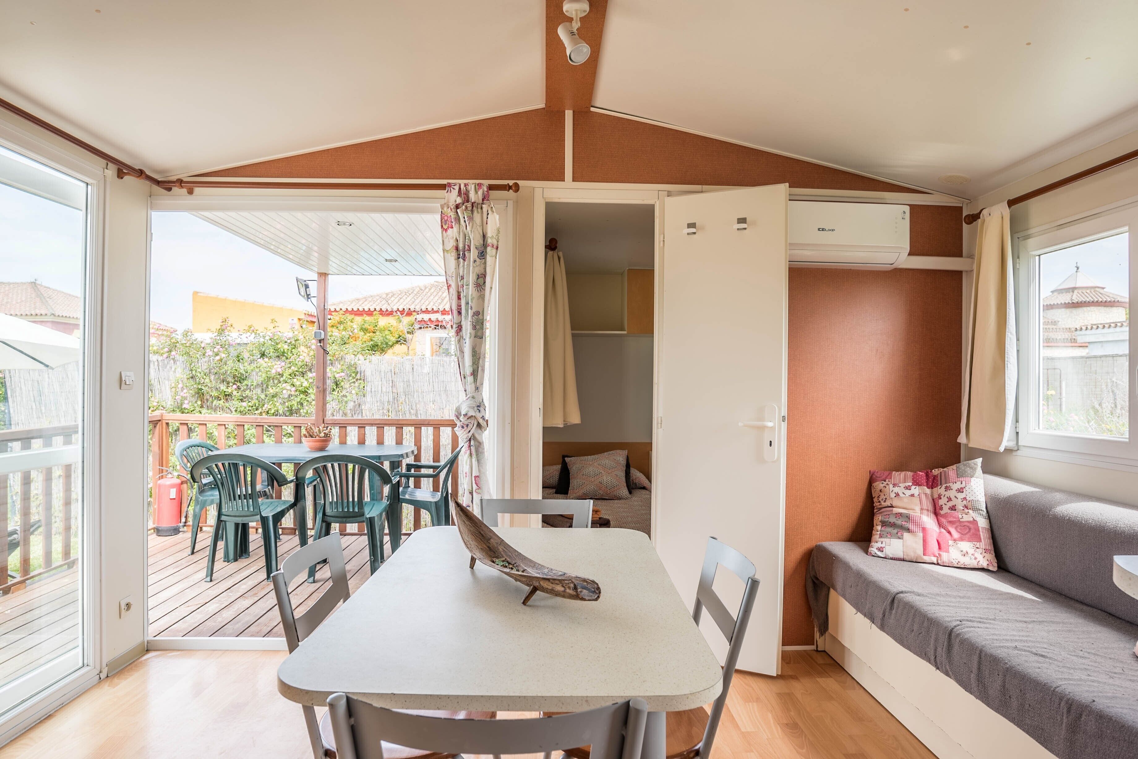 Una habitación con una mesa, sillas, un sofá, un ventanal y una puerta abierta que da a un espacio exterior con terraza y sillas.