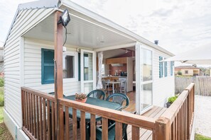 Une maison mobile dotée d’une terrasse en bois, d’un coin repas avec des chaises et une table, et d’une cuisine visible à travers une porte ouverte.