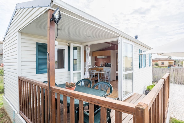A mobile home with a wooden deck, a dining area with chairs and a table, and a kitchen visible through an open door.