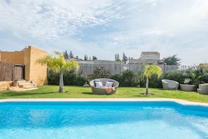 A swimming pool with a lounge chair and a small table, surrounded by a wooden fence and greenery.