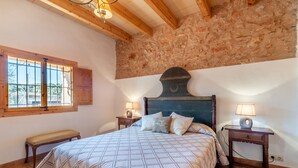 A bedroom with a large bed, wooden headboard, two bedside tables with lamps, a bench, and a stone wall. - Charming Holiday Home Can Bonjesus with Mountain View, Wi-Fi, Garden & Terrace; Parking Available, Pets Allowed (Felanitx)