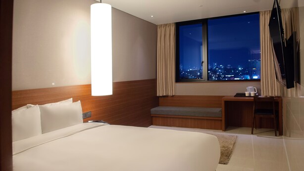 A hotel room with a large bed, a desk, a chair, a TV, and a window with a city view. - Top Cloud Hotel Cheonan (Cheonan)