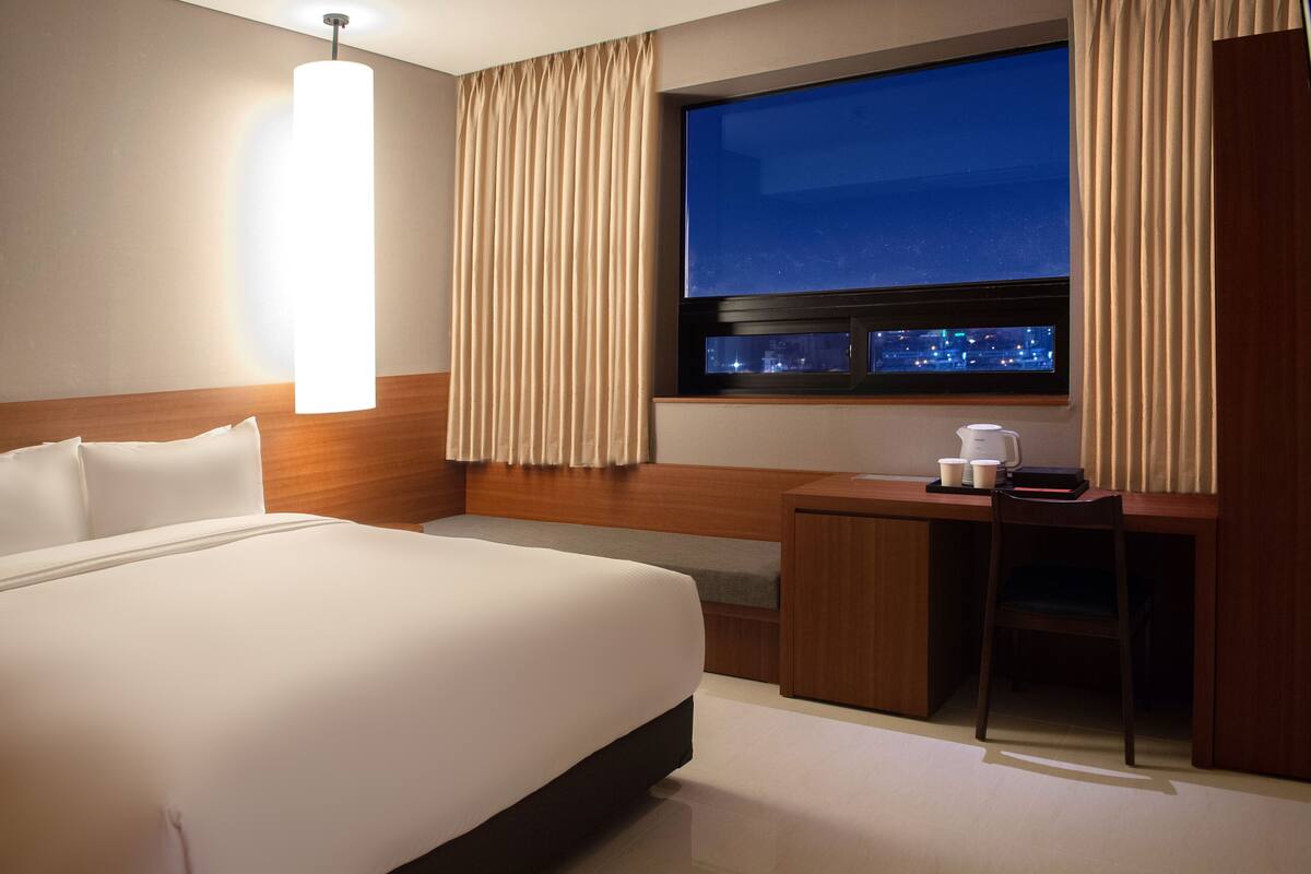 Habitación de hotel con una cama grande, un escritorio con silla, cafetera y ventana con cortinas.