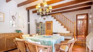 Un coin repas avec des chaises en osier, une table recouverte d’une nappe à rayures, un lustre, un escalier en bois et un buffet aux portes vitrées.