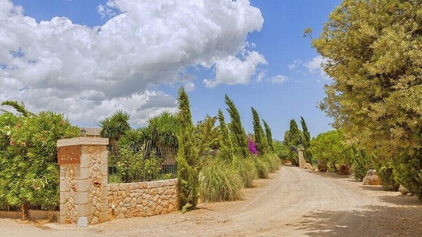 Un chemin de campagne menant à une porte en pierre surmontée d’une enseigne, le tout entouré d’une végétation luxuriante et d’arbres.
