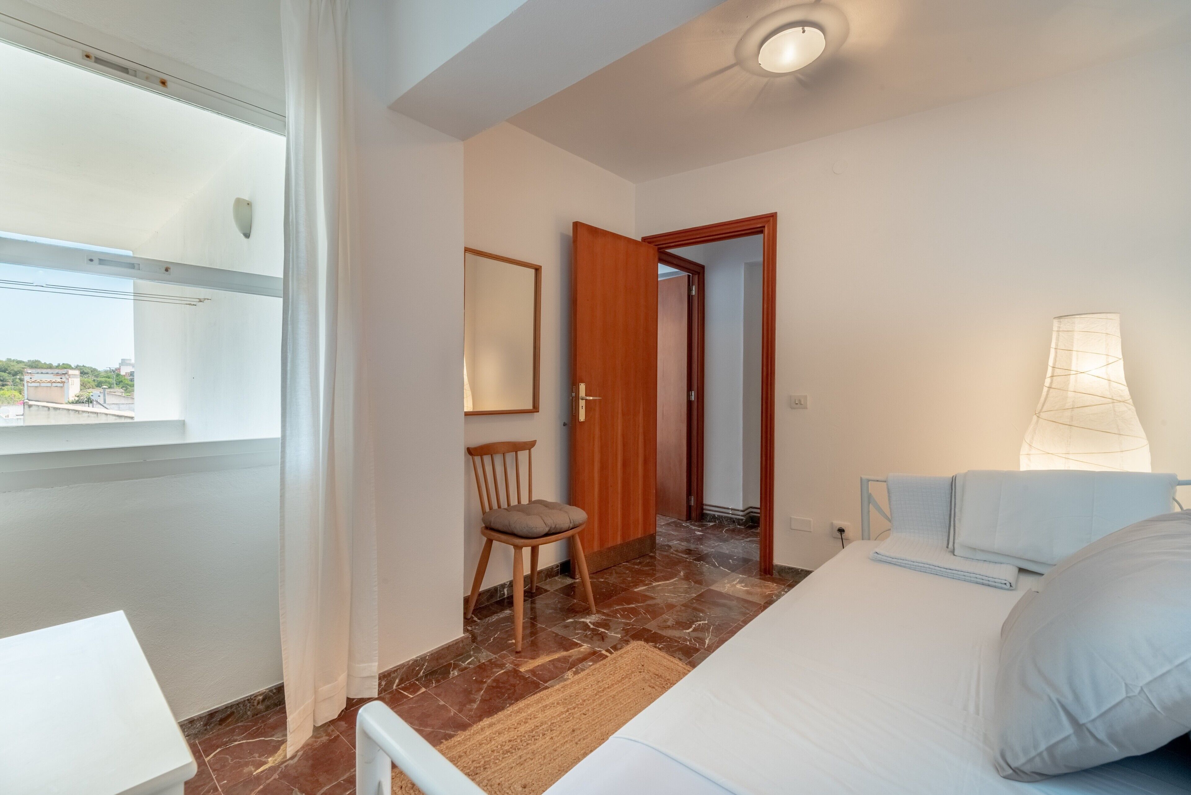 Un dormitorio con cama, una silla, un escritorio, una lámpara y vistas al exterior.