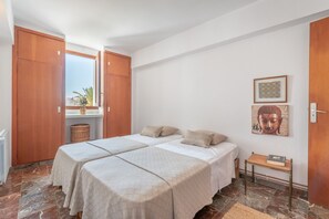 A bedroom with two beds, a window with a view, a small table with a book, and a wall with a framed picture.