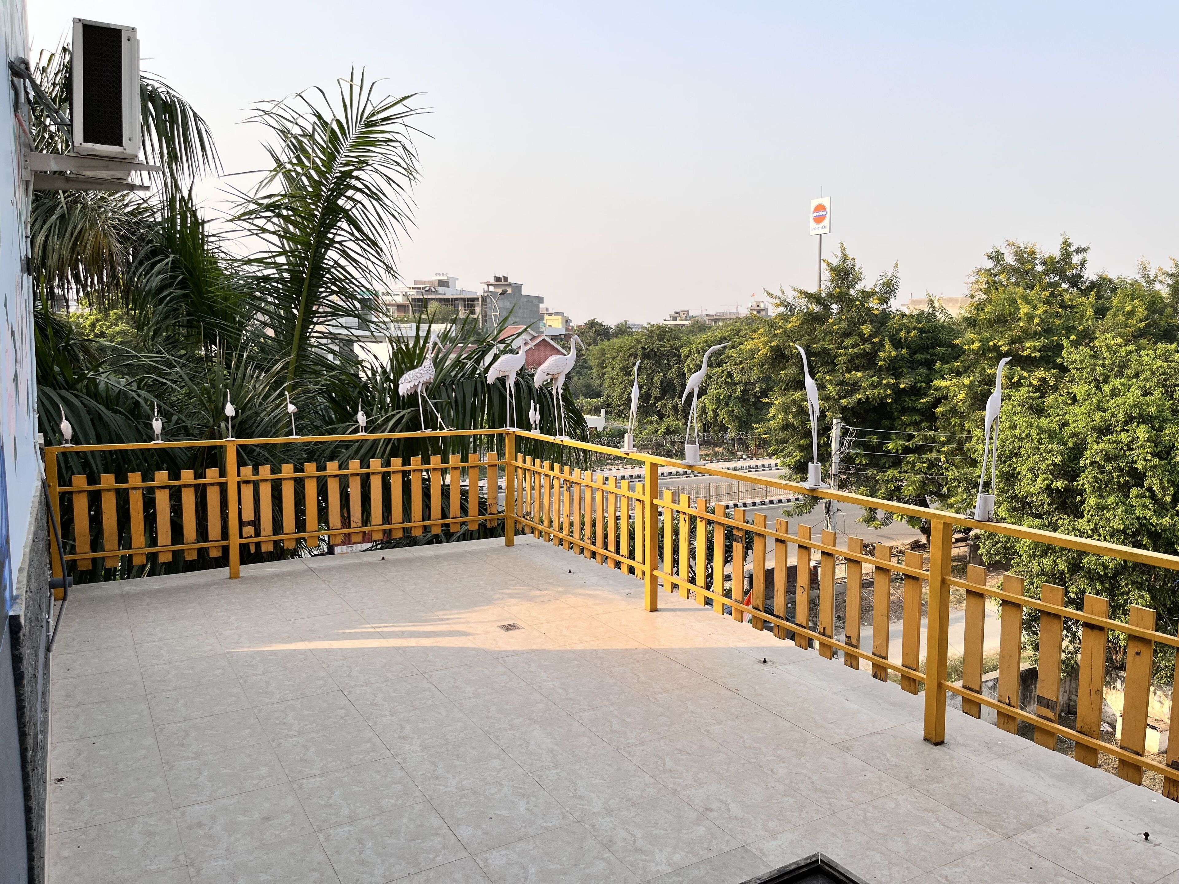 A terrace with a yellow railing, surrounded by greenery and buildings.