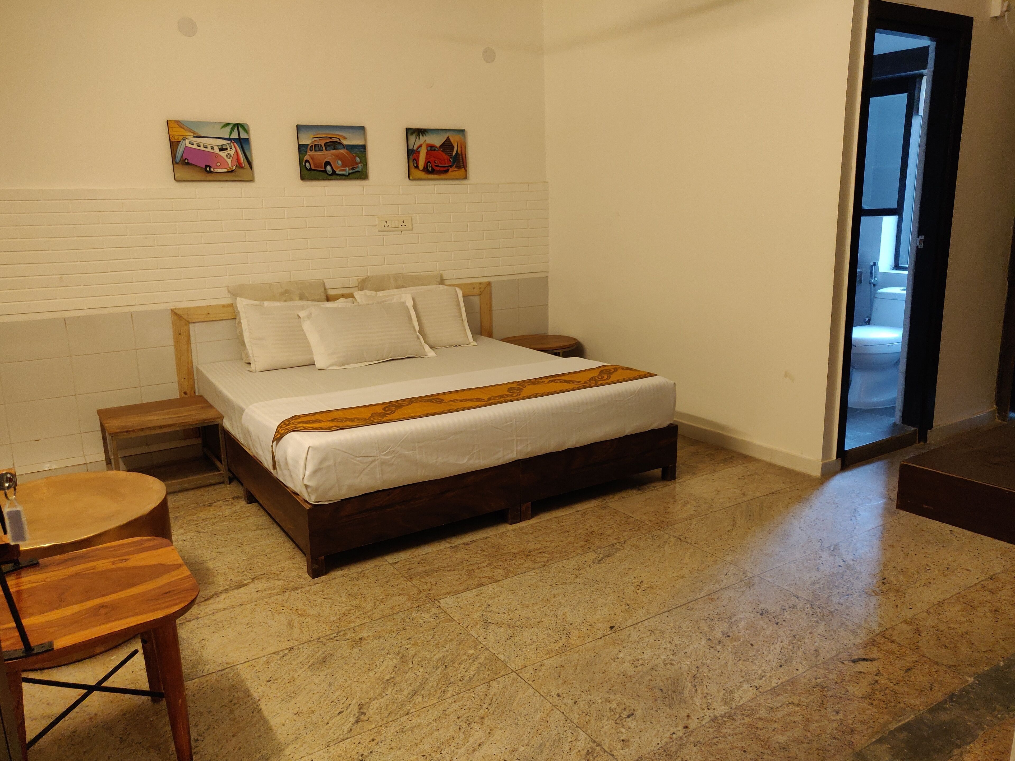 Un dormitorio con cama, cabecera de madera, paredes blancas y suelo de baldosas. Hay tres cuadros en la pared y una puerta que da al baño.