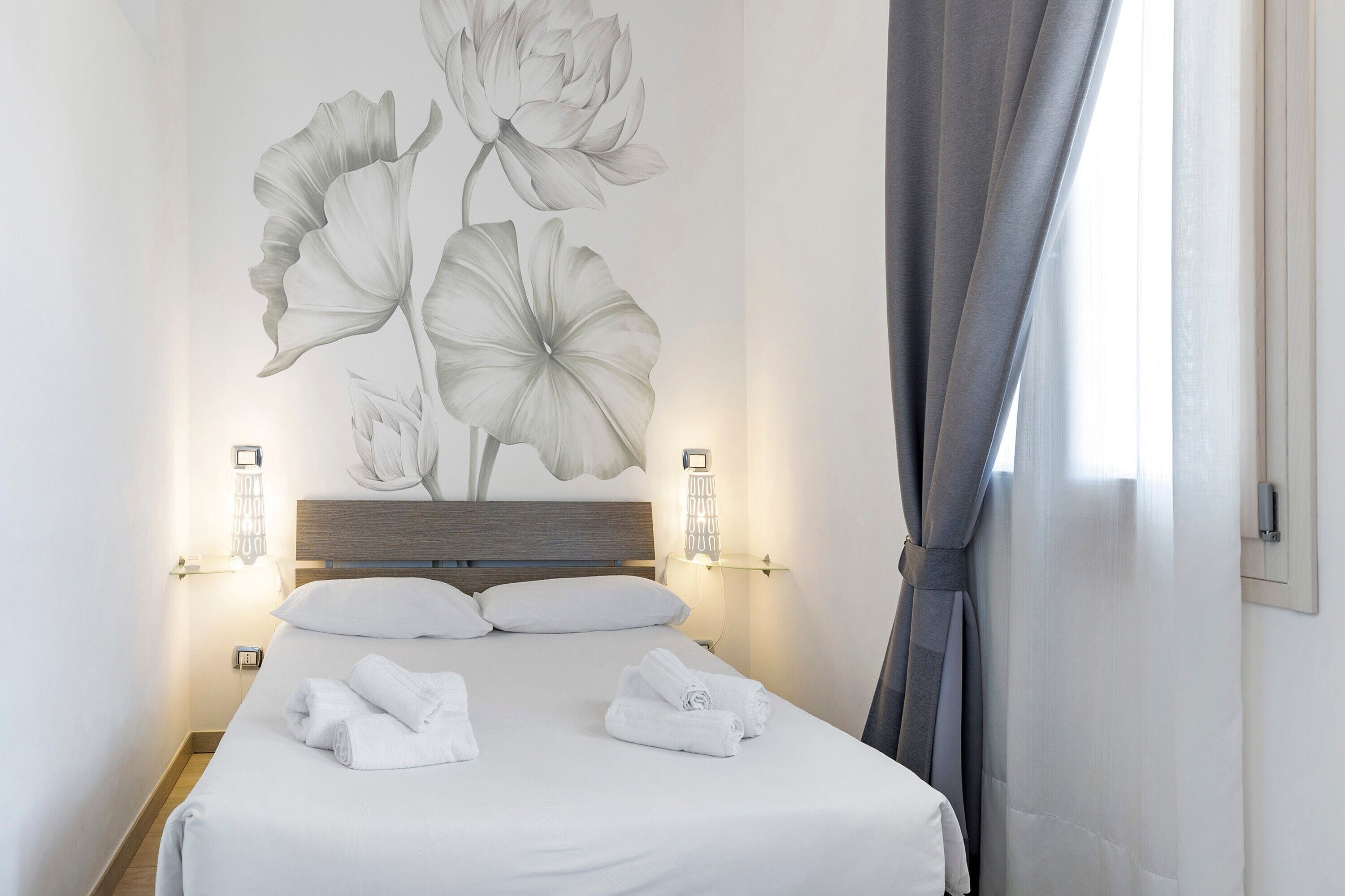 A hotel room with a bed, white linens, a large floral wall art, two bedside lamps, and a window with curtains.