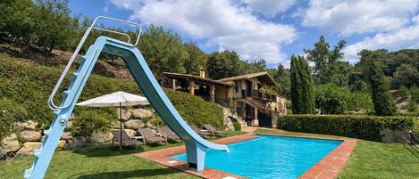 Una piscina con un tobogán azul, rodeada por un césped cuidado y un muro de piedra.