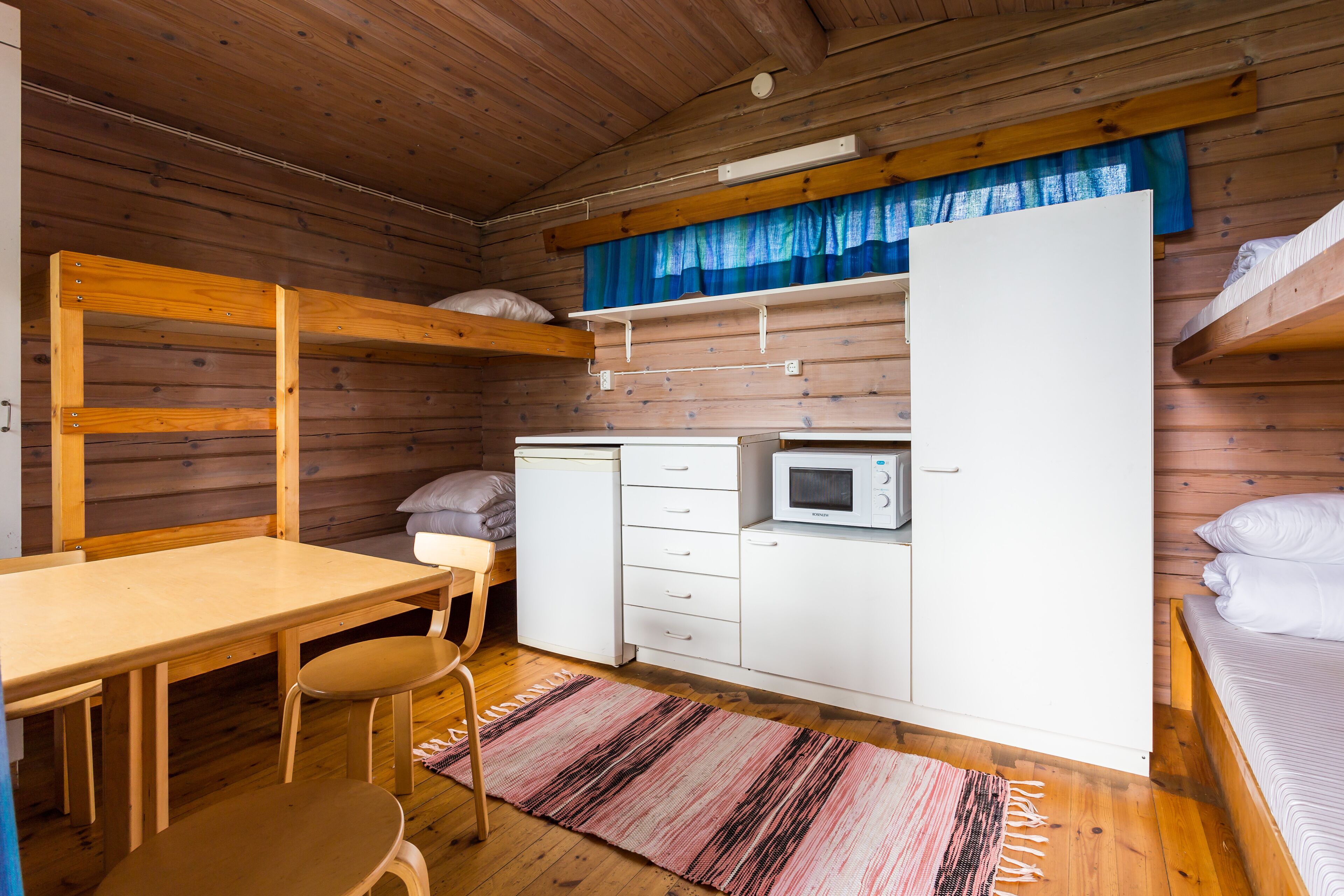A compact room with bunk beds, a small kitchenette, a wooden table with two chairs, and a striped rug.